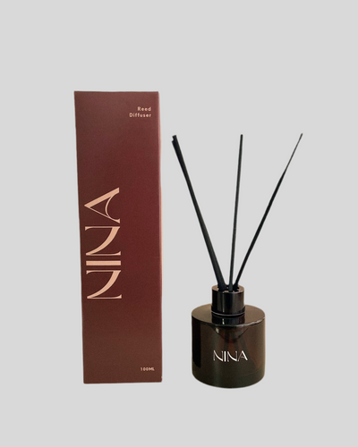 Afbeelding van een Nina Studio geurstokjes diffuser met een modern en minimalistisch ontwerp. De doos heeft een dieprode kleur met de merknaam “NINA” in grote, verticale letters en de tekst “Reed Diffuser” samen met de inhoud van 100ml. Naast de verpakking staat het cilindervormige diffuserflesje, gemaakt van donker glas, met het logo NINA in witte letters. Drie zwarte stokjes steken stijlvol uit het flesje, wat een elegante uitstraling geeft. De geur van de diffuser is “Bessen” 