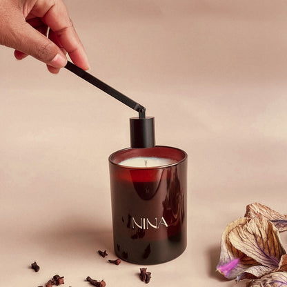 Hand steekt een zwarte metalen kaarsendover in een luxe geurkaars met roodbruine glazen houder met het logo 'NINA'. De scène wordt omlijst door gedroogde bloemen en kruidnagels, die samen een warme, elegante sfeer uitstralen.